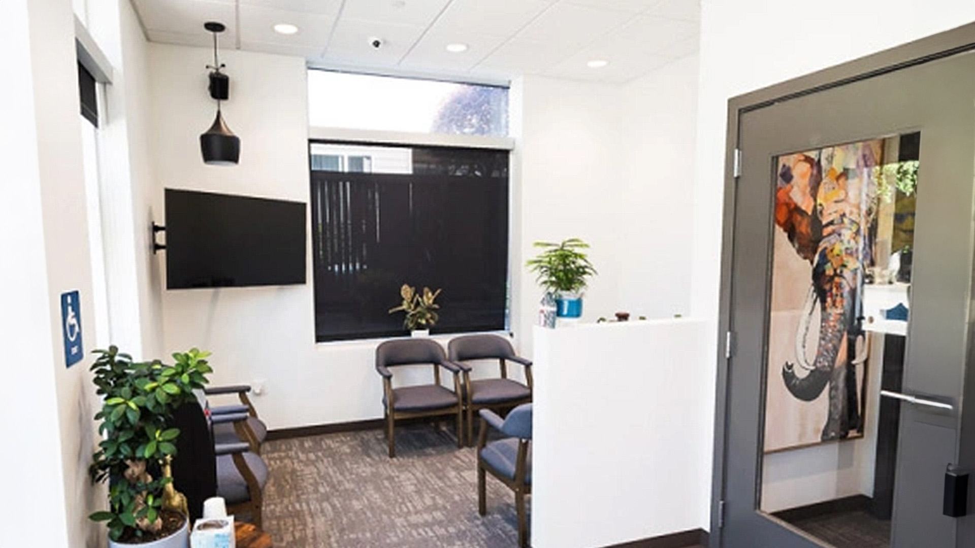 The image shows an interior view of a dental office waiting room with a modern design, featuring a reception desk, chairs, artwork on the wall, a window, and a potted plant, all under natural light.
