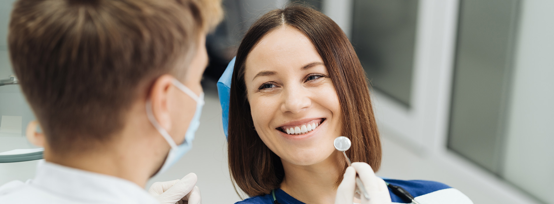 The image shows two individuals engaged in a dental appointment with one person seated in a dentist s chair receiving care while another stands, likely a dental professional, wearing a mask and using a dental tool.