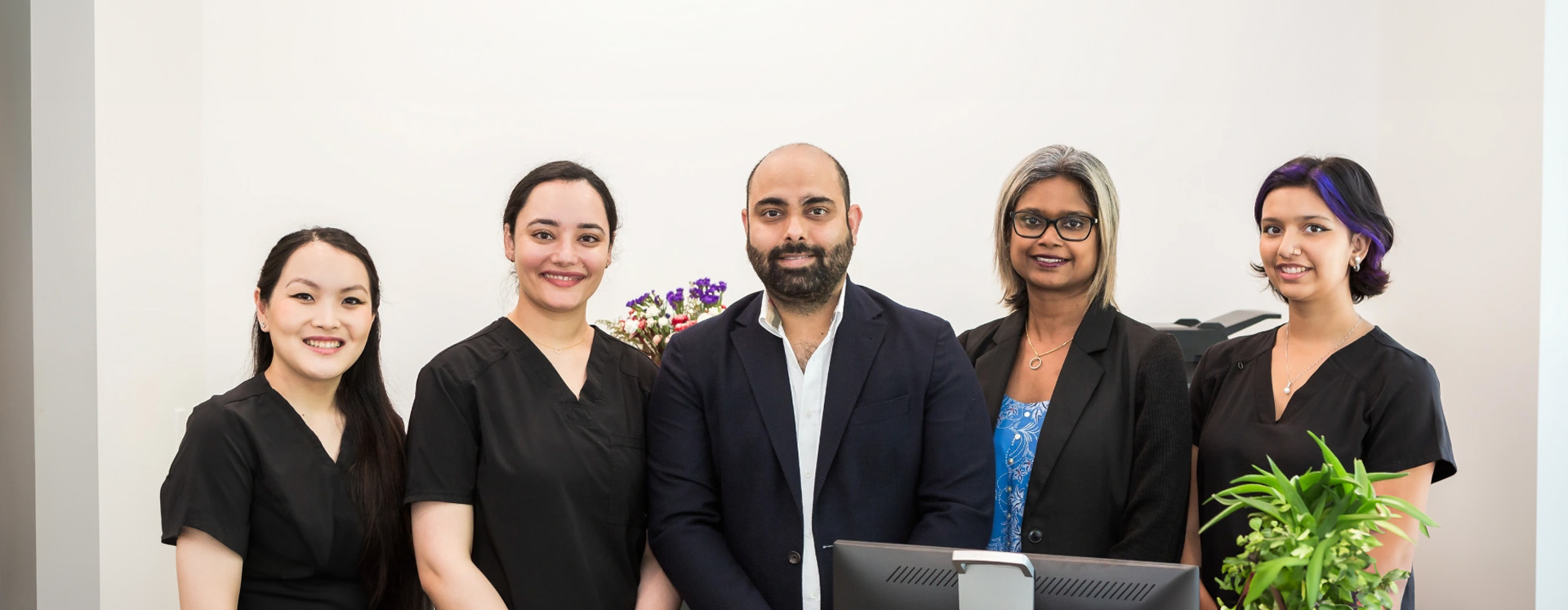 The image shows a group of six individuals posing together for a photo they appear to be professionals, possibly healthcare workers, standing in front of an office environment with a smiling receptionist behind them.