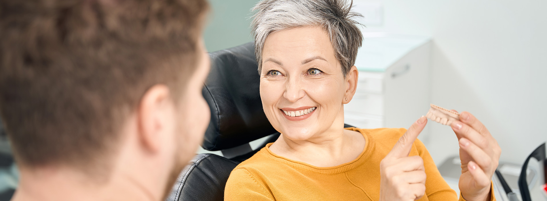 The image shows a woman sitting in front of a man, both looking towards each other with smiles on their faces, in an indoor setting with a blurred background.