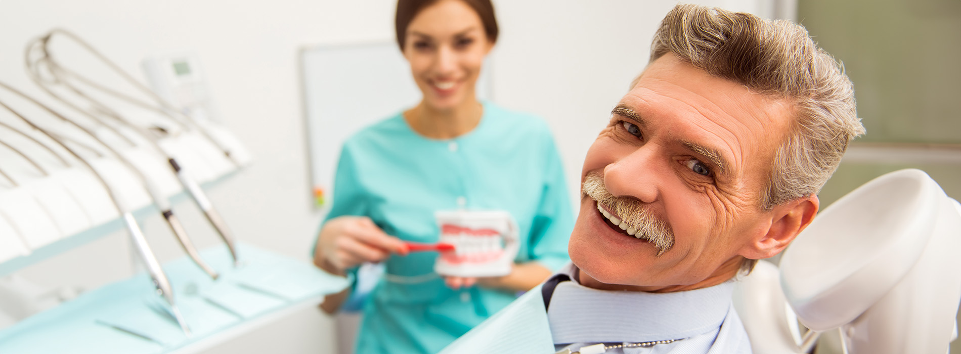 The image shows two people in a dental office setting a man with a mustache holding a red object and smiling, standing next to a woman wearing scrubs who seems to be presenting something to him, while there are various dental equipment in the background.