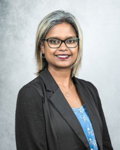 The image shows a person standing against a background with a neutral expression, wearing glasses, a dark jacket over a light top, and has short hair.