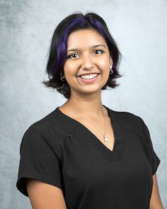 The image features a person posing for a professional portrait with a smile. They are wearing a dark shirt, have short hair, and appear to be standing against a neutral background.