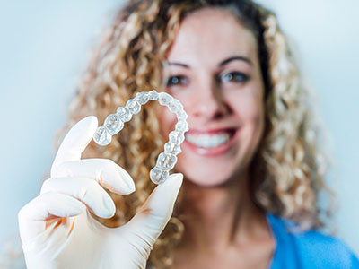 A woman holds up a clear plastic tray containing a set of transparent teeth-shaped objects against her mouth.