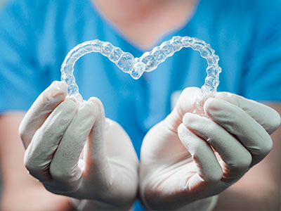 A hand holding a heart-shaped clear plastic object, possibly a dental appliance, against a blue background.