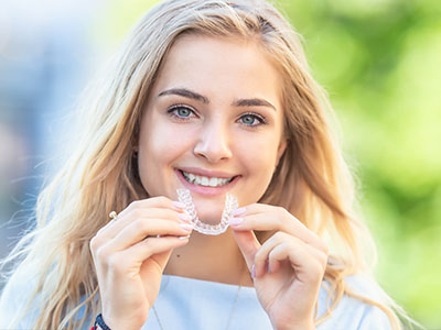 The image shows a young woman with blonde hair smiling at the camera while holding a toothbrush in her mouth.