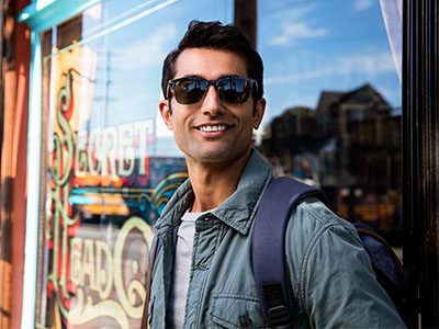 The image features a man standing in front of a storefront window, wearing sunglasses, a backpack, and a jacket, with a smile on his face.