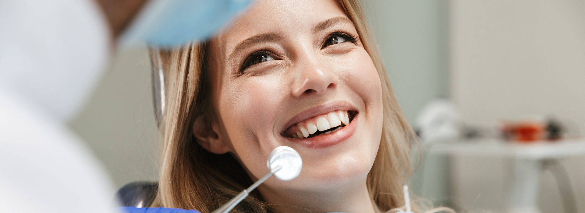 A woman with a big smile sitting in a dental chair, being attended to by a dentist.
