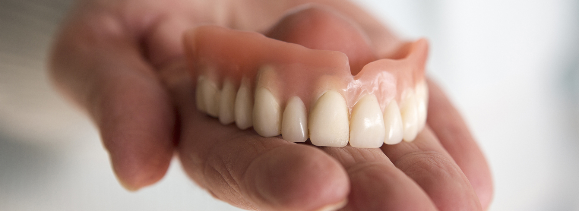The image shows a person s hand holding an upper denture with multiple missing teeth.