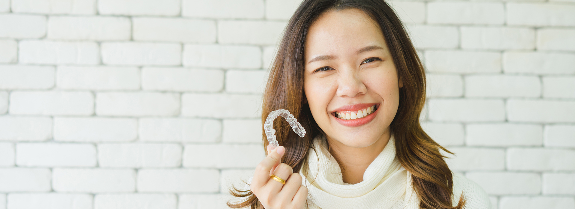 The image features a smiling woman holding up a toothbrush with her right hand against a brick wall background.