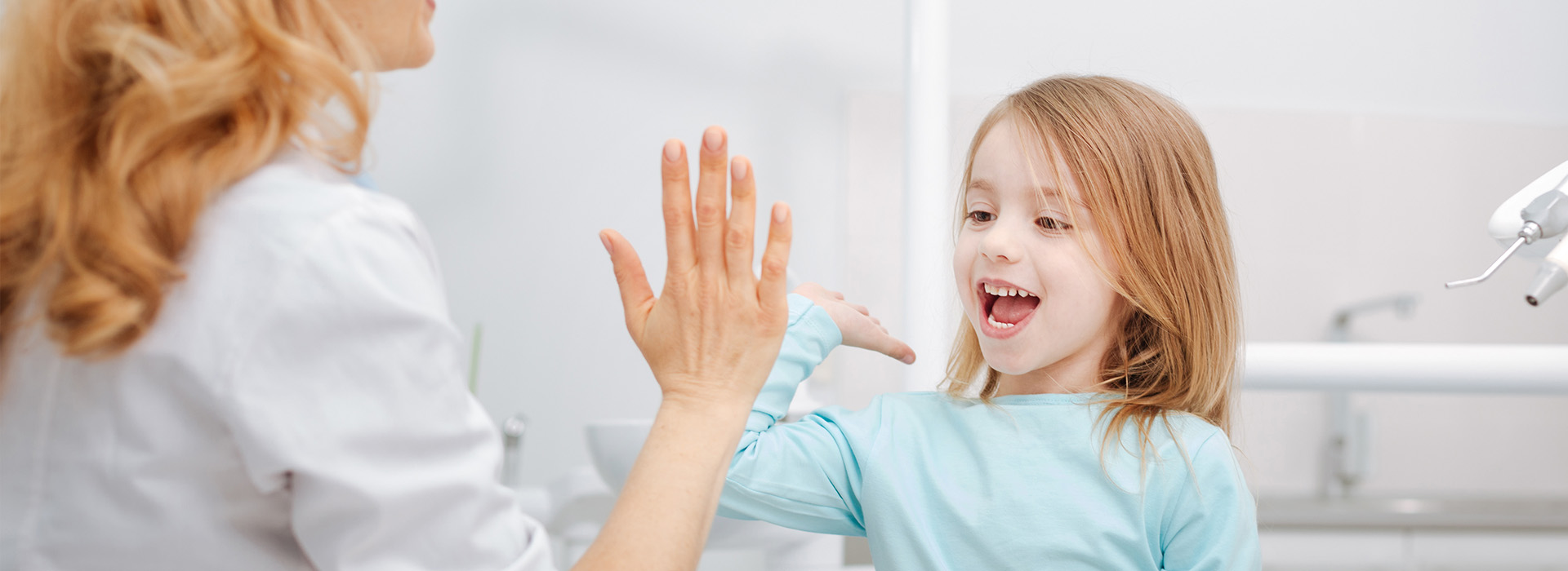 The image shows a young girl with blonde hair, wearing a blue shirt, waving at a woman who is smiling and holding up her hand towards the camera. They are indoors, likely in a bathroom given the visible sink and mirror.