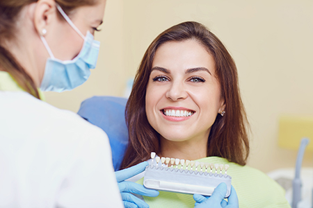 A woman with a smile sits in a dental chair, receiving a teeth cleaning from a professional who uses an electric toothbrush.