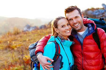 The image depicts a man and a woman posing together outdoors during daylight, with a backdrop of natural scenery, both wearing backpacks and smiling.