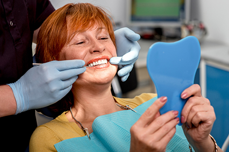A woman sitting in a dental chair, smiling broadly, with dental tools around her neck, holding up a blue toothbrush-shaped object, while another person applies a dental impression material on her teeth.