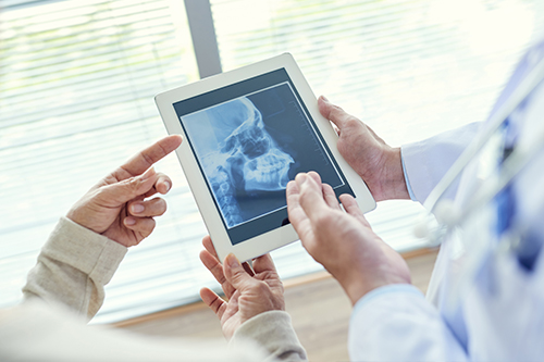 A healthcare professional showing a patient an X-ray on a tablet with a large monitor displaying a medical image.