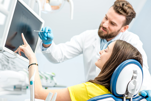 The image shows a person sitting in a dental chair with a dental professional standing next to them, both are wearing medical gloves and face masks, indicating a dental examination or treatment taking place during the COVID-19 pandemic.
